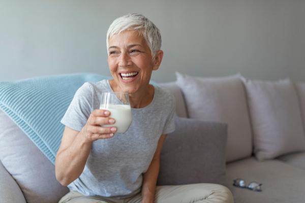 Une femme blanche aux cheveux blancs courts est assise sur un canapé en souriant, tenant un verre de lait.