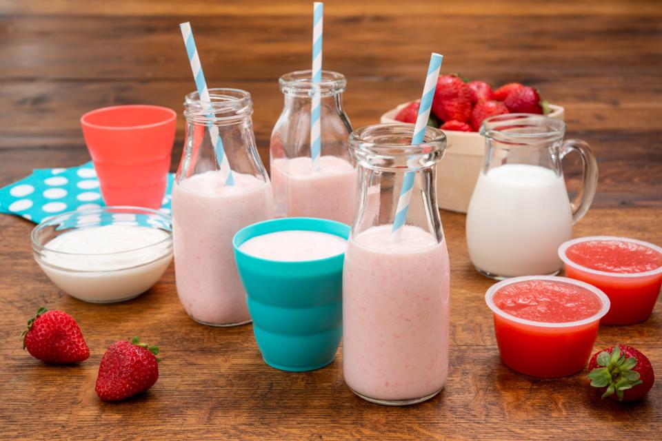 Smoothie aux fraises dans des bouteilles de verres et des petits verres avec des pailles sur une table. Il y a des fraises fraîches et des coupes de sauce aux fruits aussi sur la table.